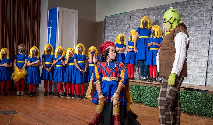 Lord Farquaad talking with Shrek surrounded by dancers in blue dresses and yellow wigs